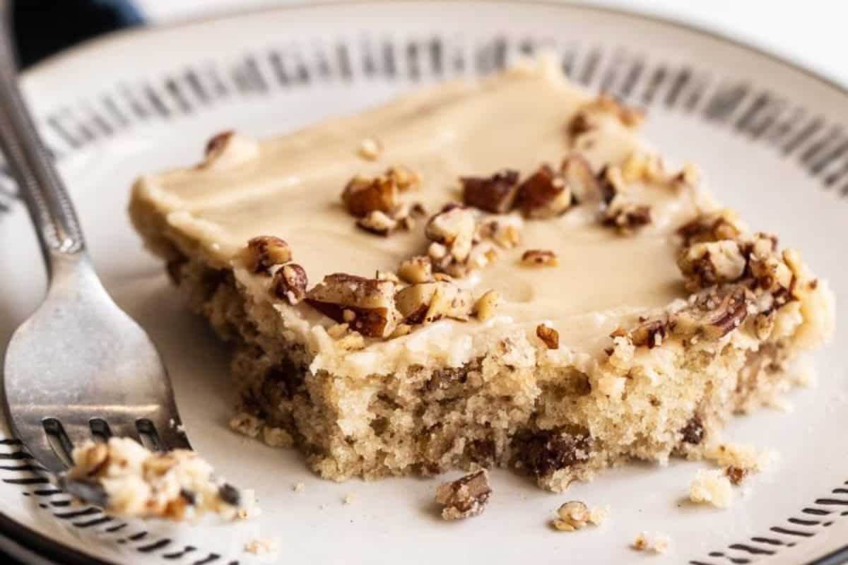 A slice of cake topped with frosting and chopped nuts is partially eaten, placed on a white plate with black detailing around the rim. A fork with cake crumbs on it rests on the side of the plate.