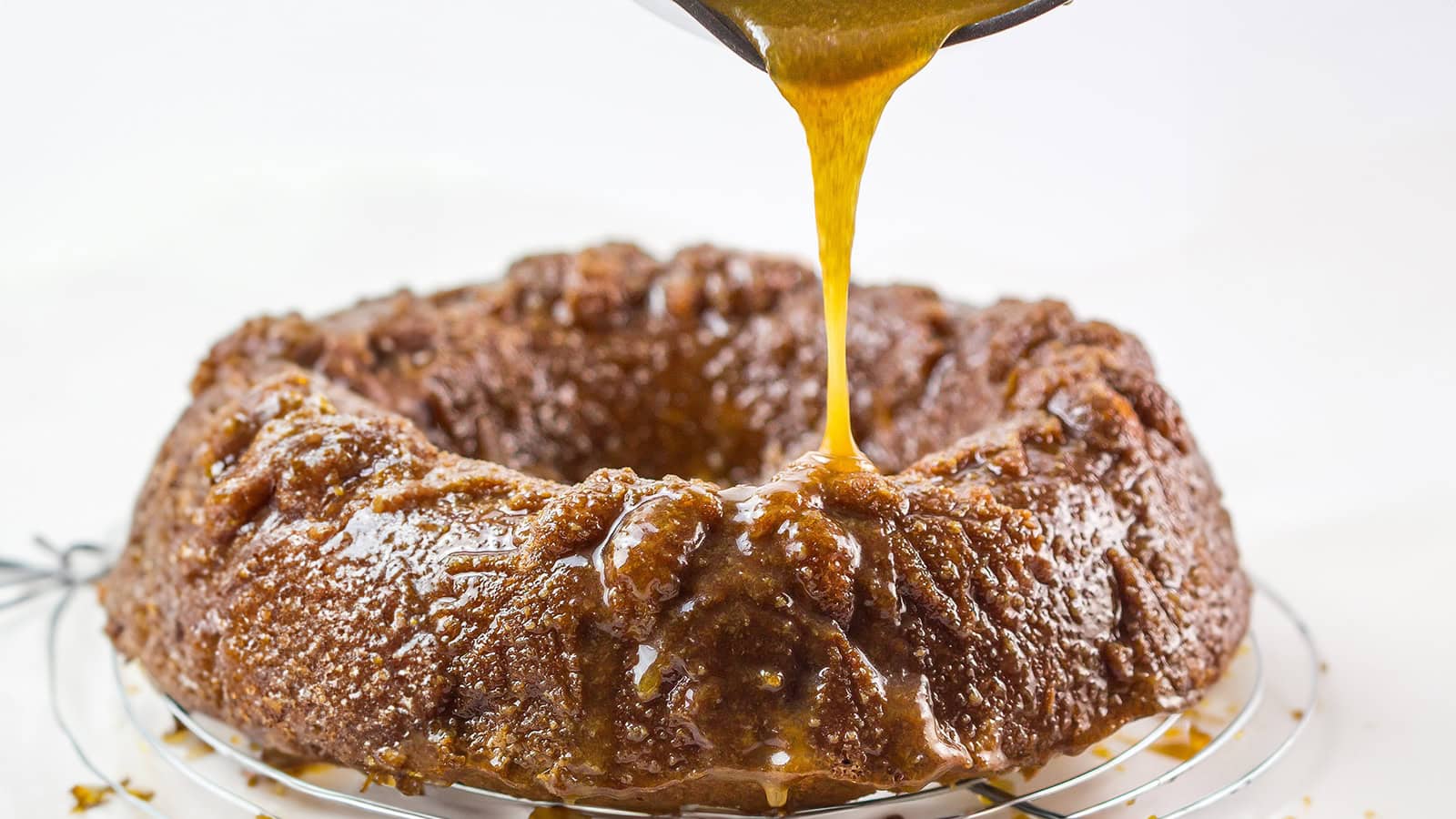 A close-up of a freshly baked bundt cake on a cooling rack. A thick, golden caramel sauce is being poured over the top of the cake, creating a glossy, rich coating. The background is softly blurred, drawing focus on the rich texture and sheen of the cake and sauce.