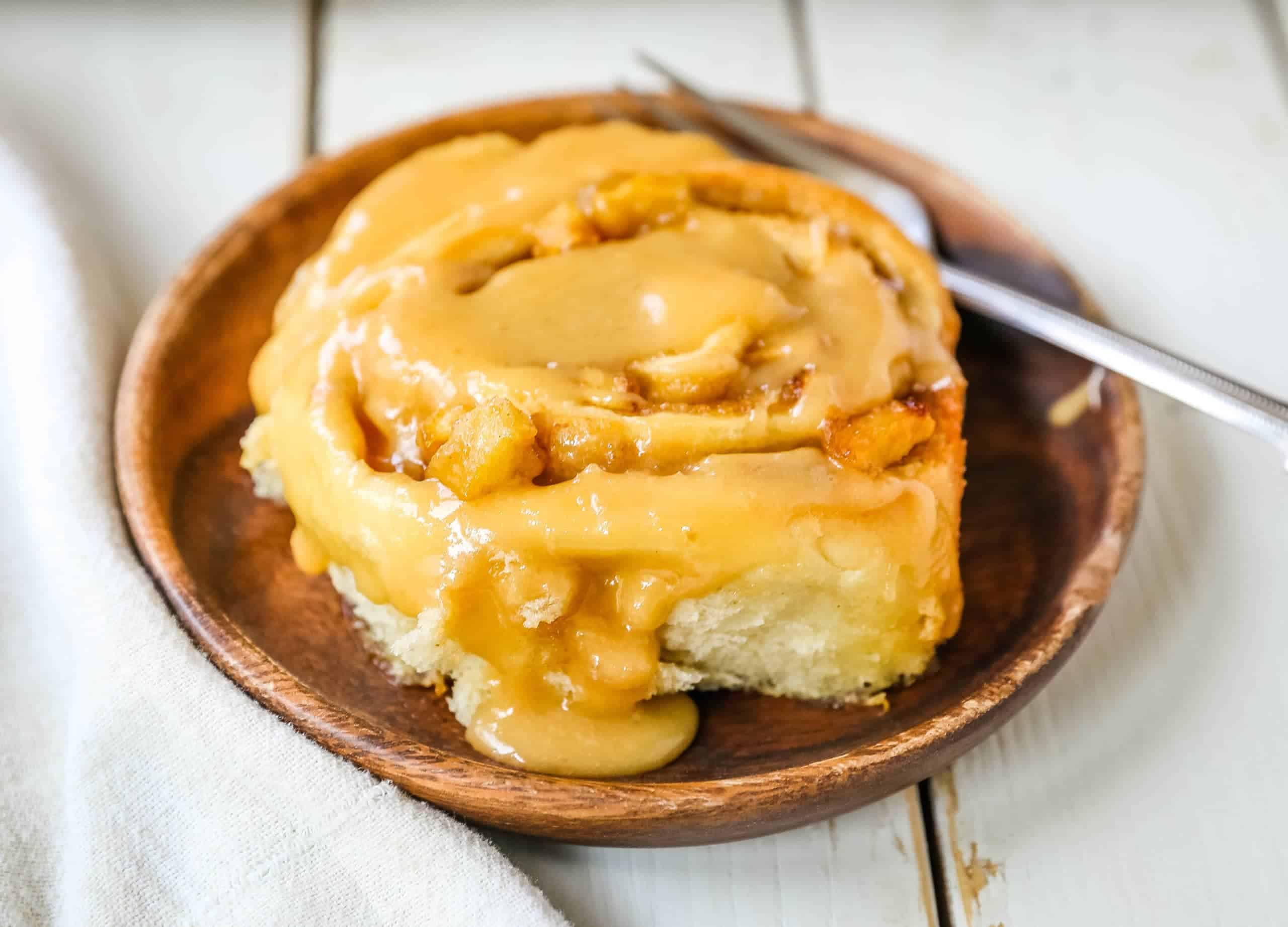 A close-up of a freshly baked cinnamon roll covered in a generous layer of caramel glaze on a brown wooden plate. A silver fork rests beside the cinnamon roll. The background features a white tablecloth and a white wooden surface.