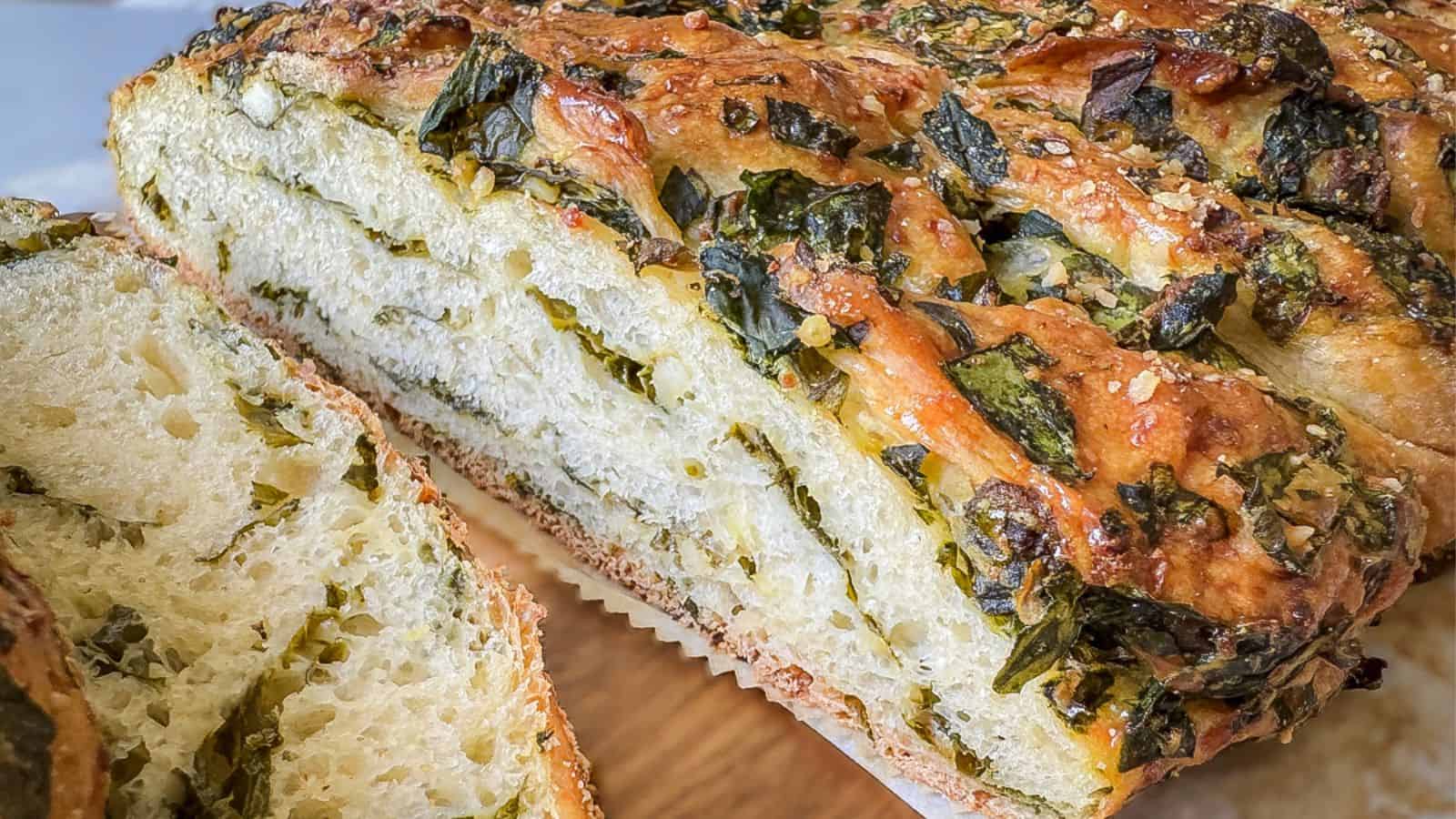 Close-up of a sliced loaf of freshly baked bread. The bread is swirled with herbs and cheese, showcasing a golden-brown crust and a soft, airy interior. The herbs are visibly green, and the cheese has a melted, slightly browned appearance.