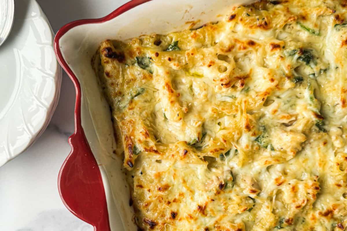 A close-up of a red-edged baking dish containing a golden-brown, baked cheesy pasta casserole with bits of green herbs visible. The dish is partially shown with a white, patterned plate beside it on a marble surface.