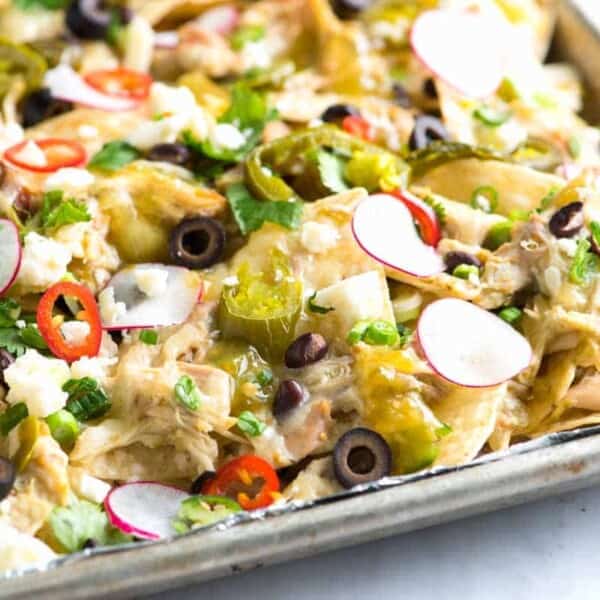 A close-up of a tray of green chicken nachos topped with various ingredients including black olives, radish slices, red chili slices, green onions, melted cheese, and cilantro. The dish appears colorful and garnished with a mix of fresh toppings.