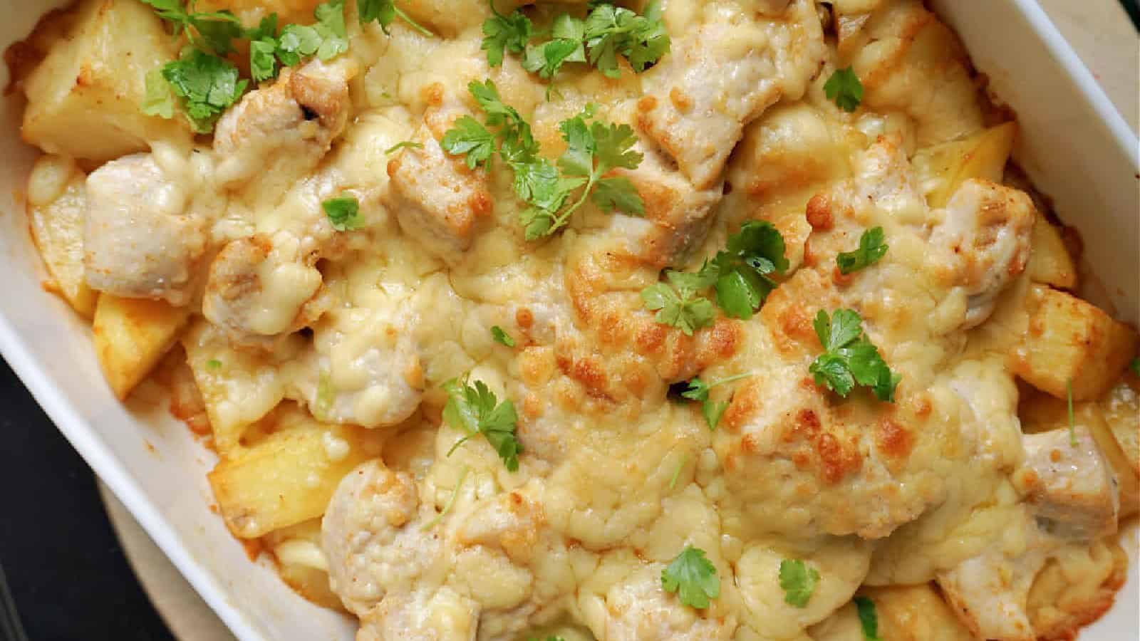 A baked casserole containing pieces of chicken, potatoes, melted cheese, and fresh parsley in a white dish.