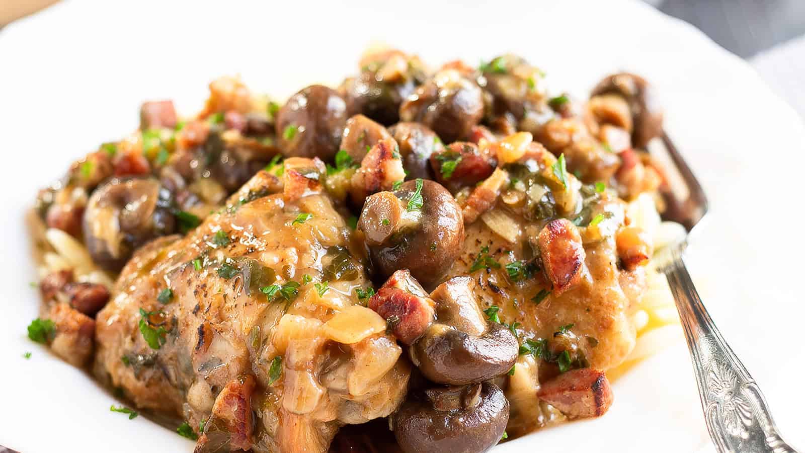 A savory dish of chicken with mushrooms, bacon, and a herb garnish served on a white plate. The ingredients are mixed in a rich, caramelized sauce, creating a hearty and appealing meal. A fork is partially visible on the right side of the plate.