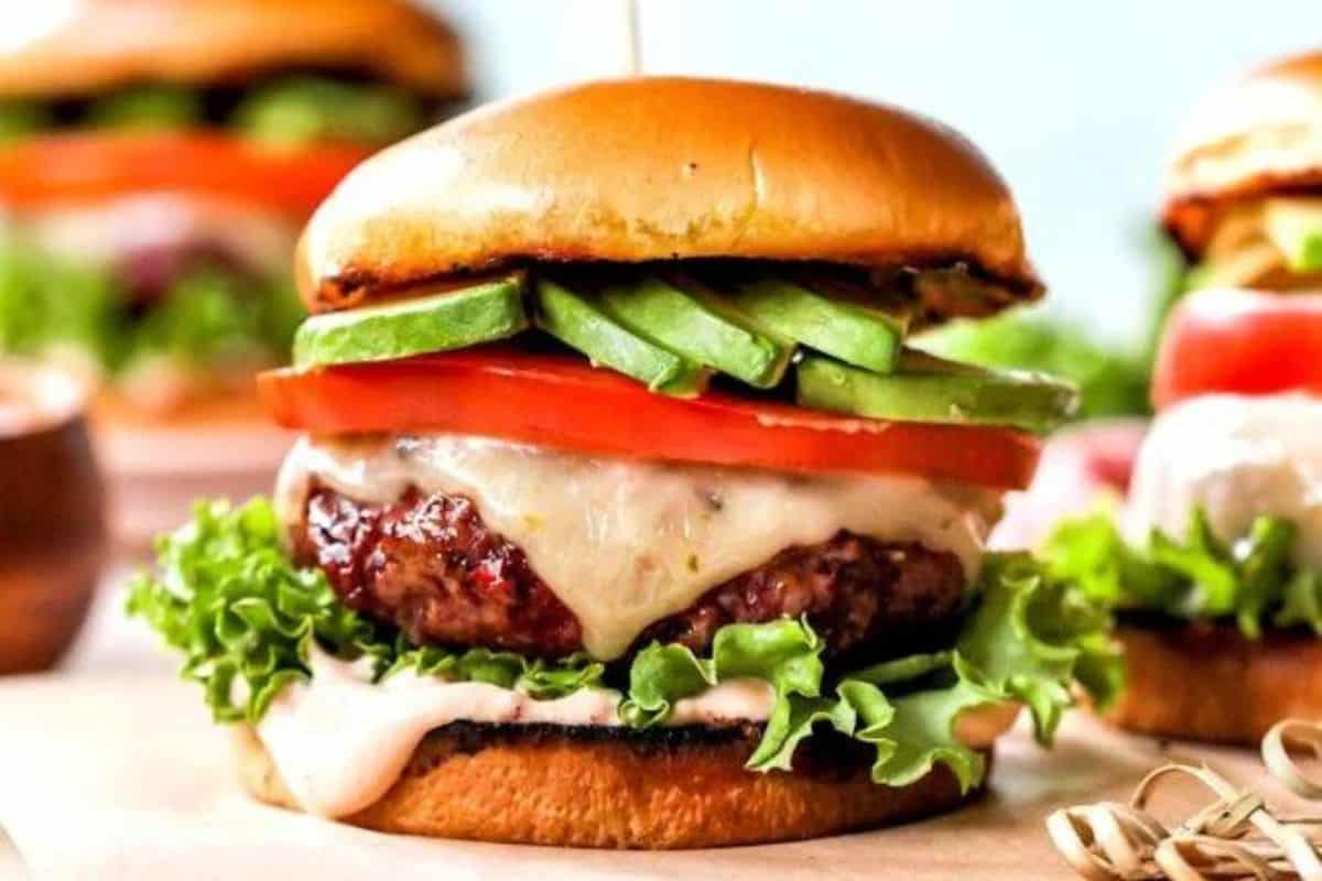 A juicy burger with a grilled patty topped with melted cheese, avocado slices, and tomato, layered with fresh lettuce on a toasted bun. One toothpick holds the burger together. Another partially visible burger is in the background.