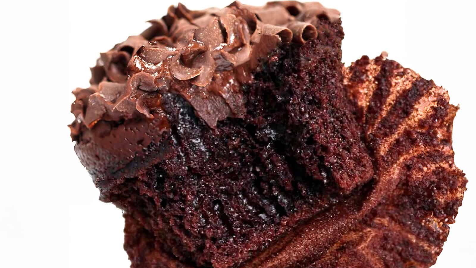 A close-up of a chocolate cupcake with rich, dark chocolate frosting. The cupcake has a bite taken out, revealing its moist interior. The cupcake liner is partially peeled away, showcasing its crumbly texture.