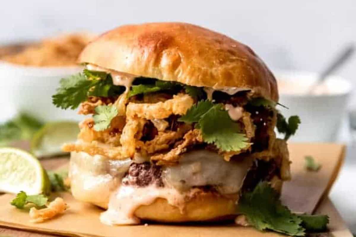 A close-up of a loaded burger with a thick beef patty, melted cheese, crispy fried onions, fresh cilantro, and a creamy sauce, all in a toasted bun. The burger is placed on a wooden board with a wedge of lime in the background.