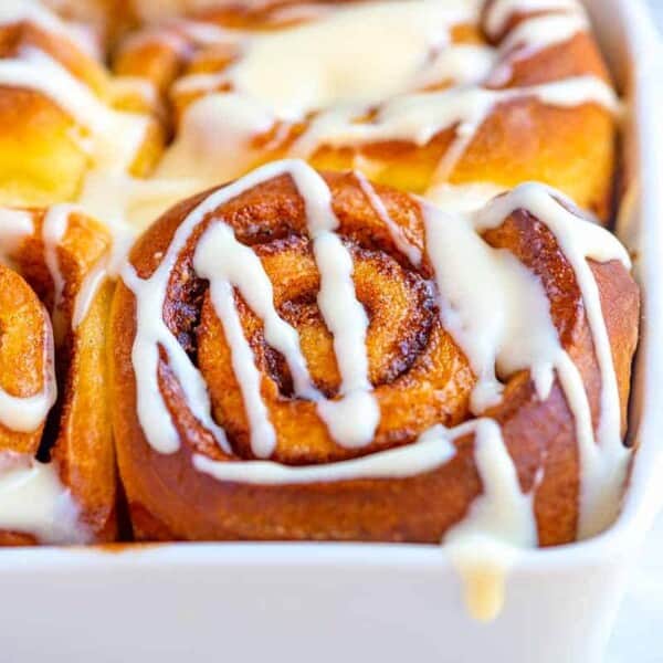 A close-up shot of a baking dish containing freshly baked cinnamon rolls drizzled with creamy white icing. The rolls are golden brown with visible layers of swirled cinnamon filling.