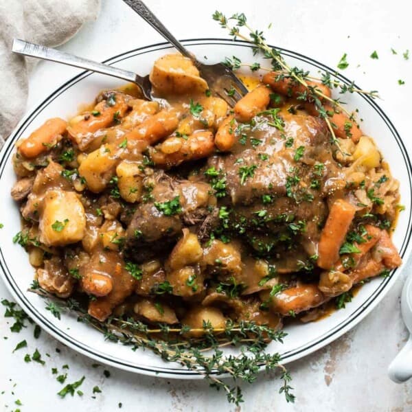 A rectangular plate filled with a hearty beef pot roast, surrounded by cooked carrots, potatoes, and fresh herbs. It is garnished with chopped parsley. A linen napkin and serving forks are to the side, with a small jug of brown gravy in the foreground.
