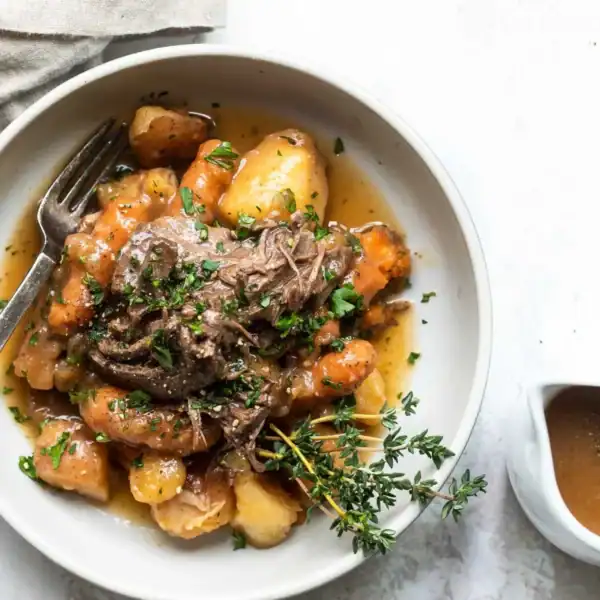 A bowl of pot roast with potatoes, carrots, and herbs sits next to a fork and a small pitcher of gravy with a sprig of thyme.