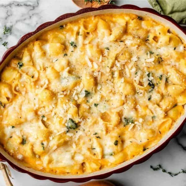 A baked pasta dish topped with melted cheese and garnished with fresh herbs in an oval ceramic baking dish. The edge of the dish is red and it is placed on a marble surface with a green cloth partially visible in the background.