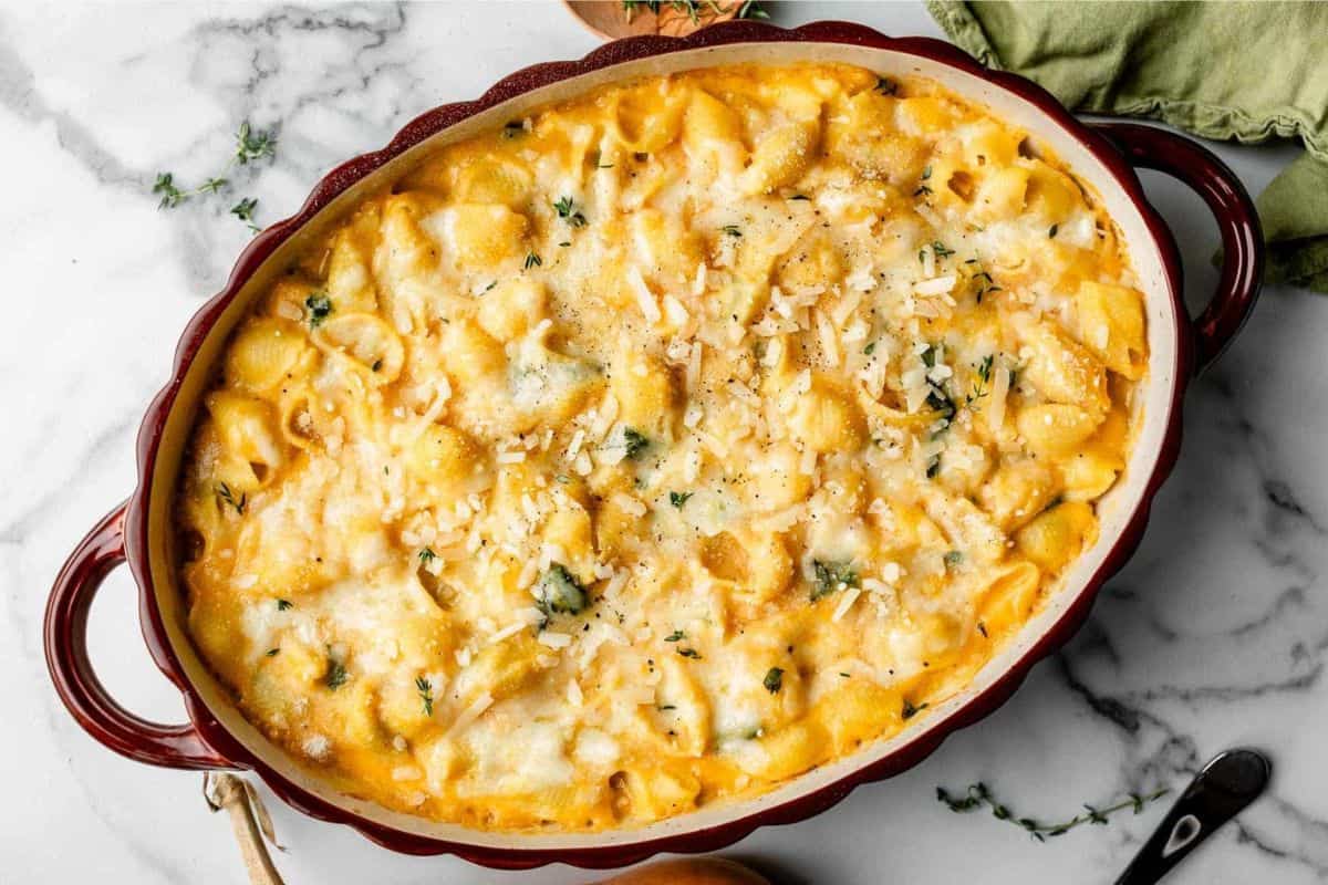 A baked pasta dish topped with melted cheese and garnished with fresh herbs in an oval ceramic baking dish. The edge of the dish is red and it is placed on a marble surface with a green cloth partially visible in the background.
