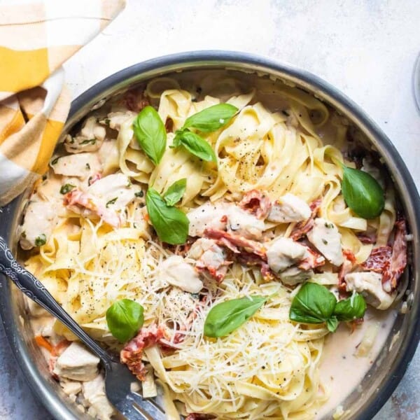 A pan of creamy fettuccine pasta with chicken, sun-dried tomatoes, basil, and grated cheese next to a yellow-checkered cloth and a glass of red wine.