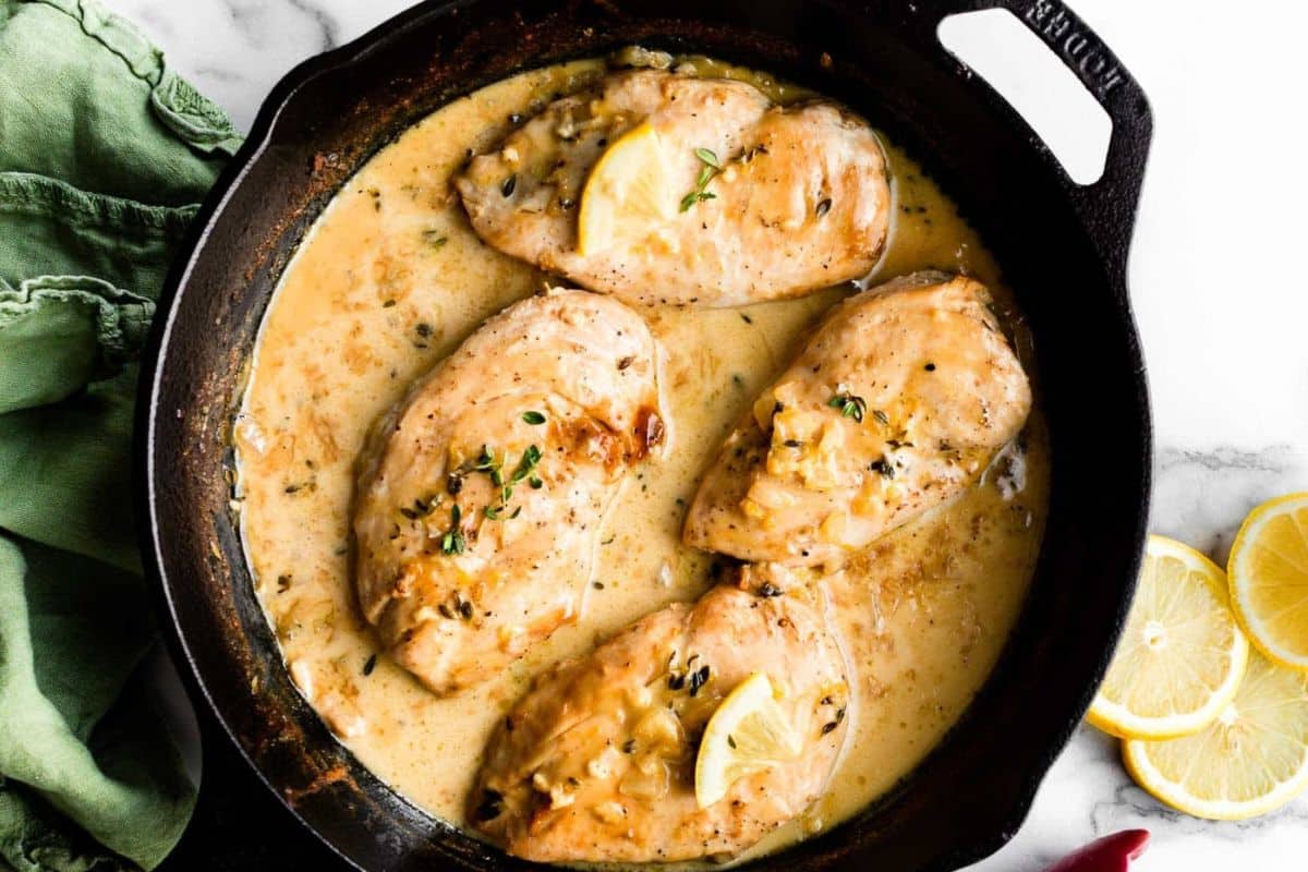 A cast-iron skillet containing four browned chicken breasts simmering in a creamy lemon-thyme sauce. Garnished with lemon slices and fresh thyme, the skillet is placed on a marble surface beside a green towel, with lemon slices on the side.