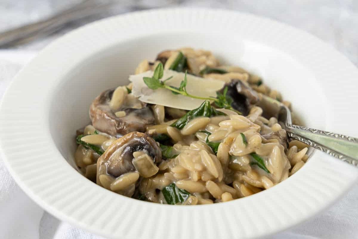 A white bowl filled with creamy orzo pasta, sautéed mushrooms, spinach, and garnished with a sprig of fresh thyme and a slice of cheese. A fork rests on the edge of the bowl.