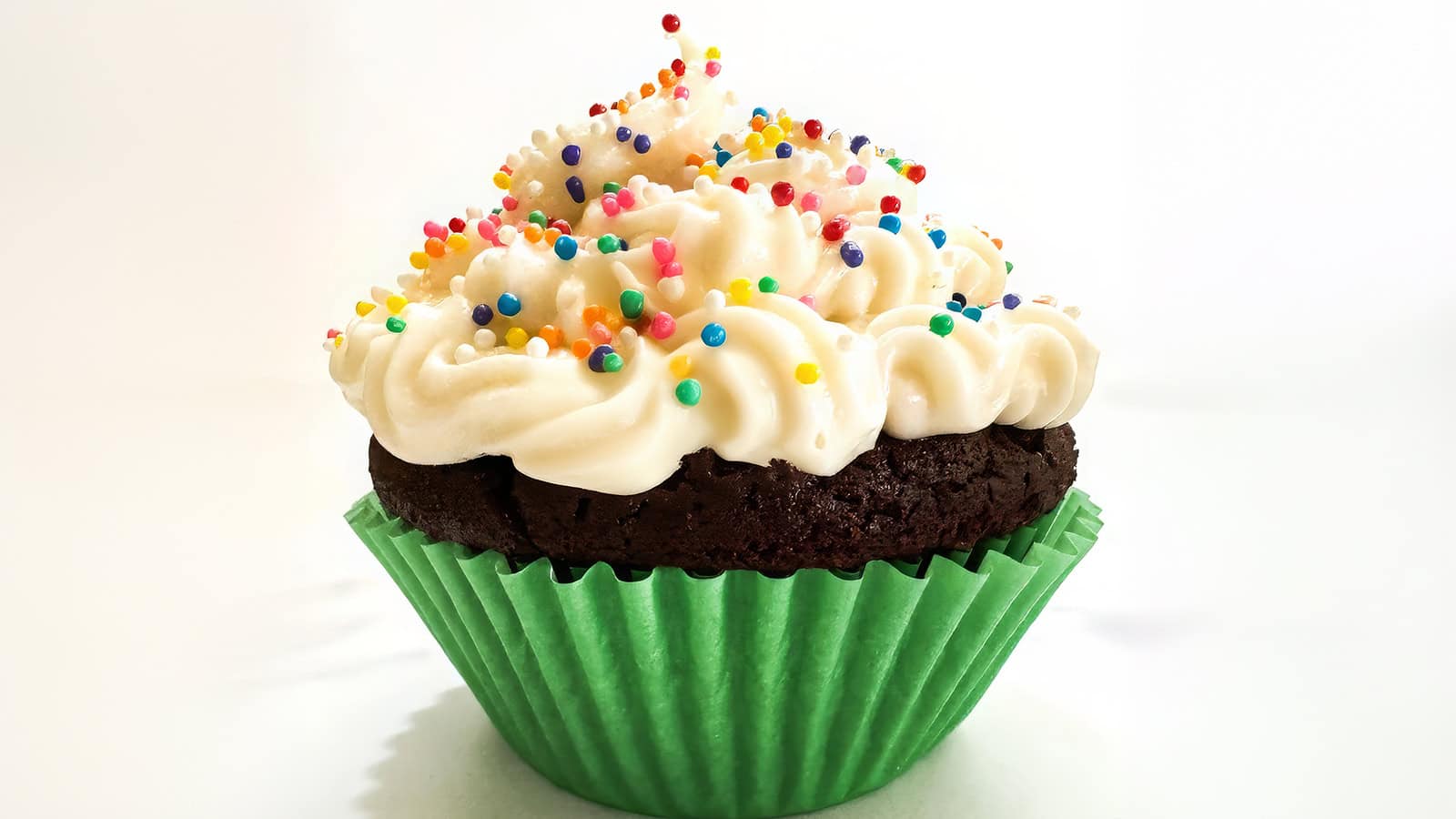 A chocolate cupcake with white frosting swirls sprinkled with colorful round sprinkles. The cupcake is wrapped in a green paper liner and is set against a white background.