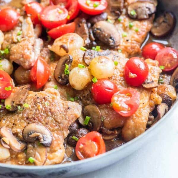 A close-up view of a skillet filled with a colorful dish consisting of cooked chicken thighs garnished with halved cherry tomatoes, mushroom slices, and pearl onions in a savory sauce. The dish is topped with finely chopped green herbs.
