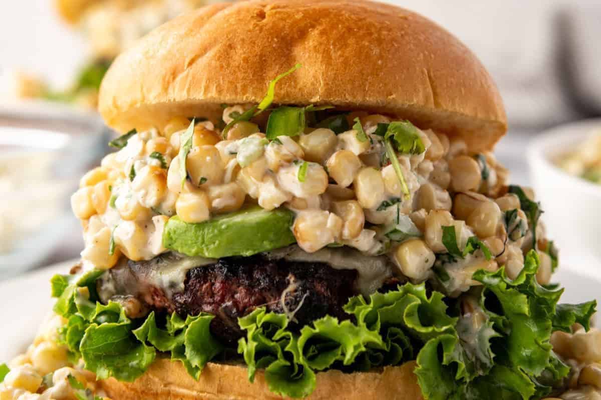 A close-up of a gourmet burger with a toasted bun, leafy lettuce, and a beef patty topped with melted cheese, avocado slices, and a generous serving of creamy, seasoned corn salad. The burger is set on a white plate with another burger seen blurred in the background.