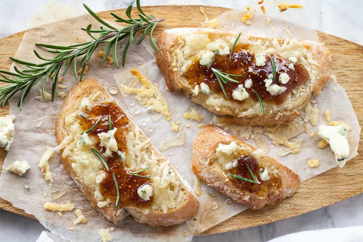 Three slices of crostini topped with melted cheese, fig jam, crumbled cheese, and rosemary are arranged on a wooden board alongside a sprig of fresh rosemary.