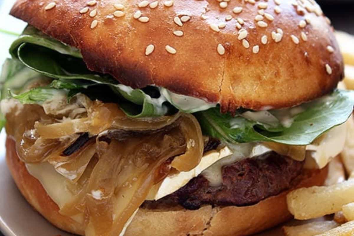 A close-up of a burger with a sesame seed bun, layered with melted cheese, caramelized onions, leafy greens, and a thick beef patty. French fries are visible beside the burger on the plate.