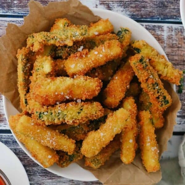 Golden-brown breaded zucchini fries are arranged on parchment paper, sprinkled with coarse salt. Sauces and fresh herbs sit nearby.