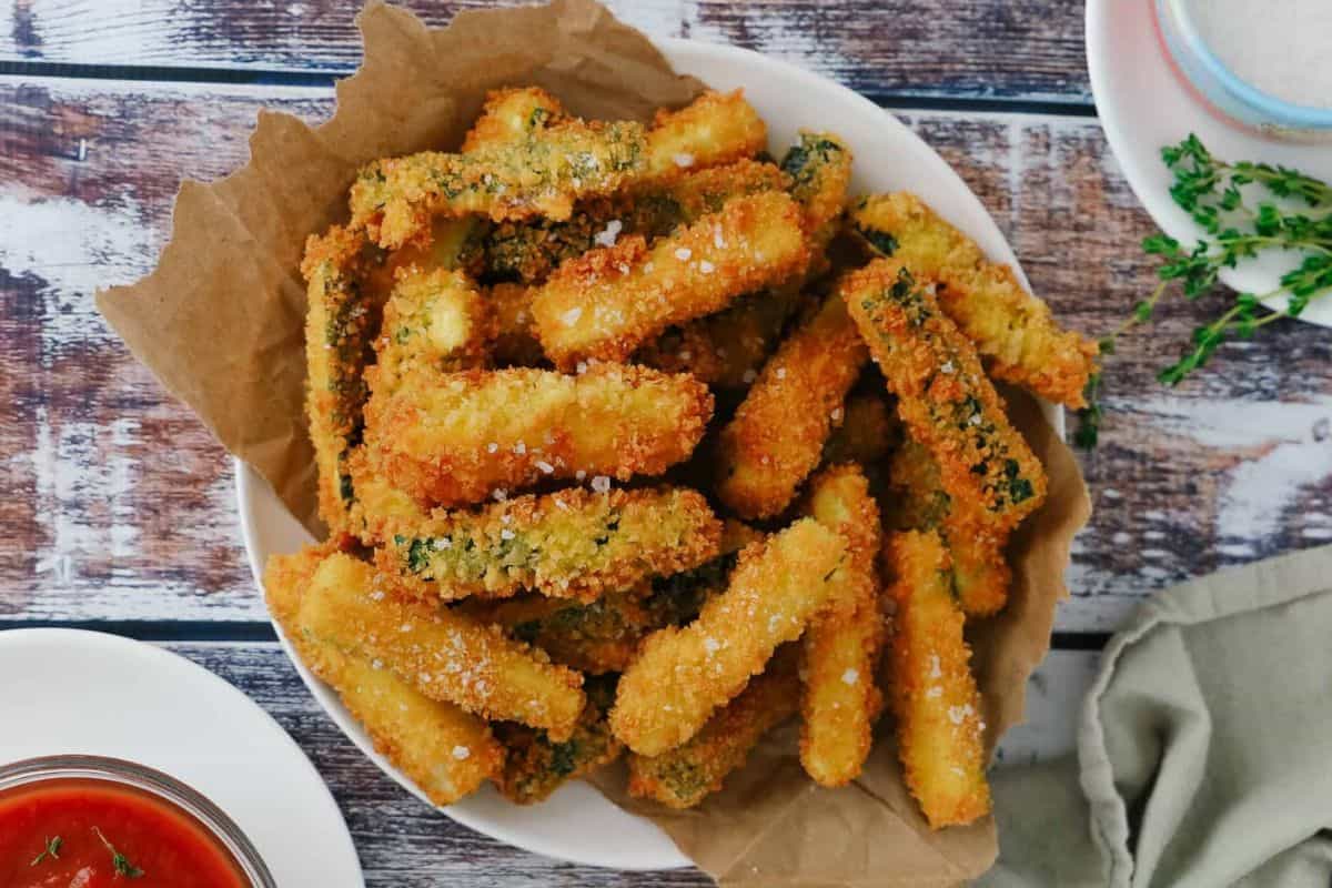 Golden-brown breaded zucchini fries are arranged on parchment paper, sprinkled with coarse salt. Sauces and fresh herbs sit nearby.