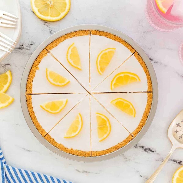 A lemon tart pie with slices, lemon wedges on top, on marble; plates, forks, lemon slices, and two pink drinks are nearby.