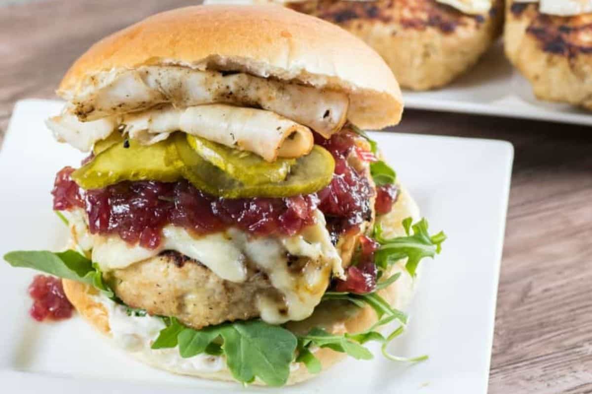 A grilled chicken burger with melted cheese, pickles, arugula, and what appears to be cranberry sauce served on a white plate. The burger is in between a toasted bun. A similarly prepared burger is visible in the background.