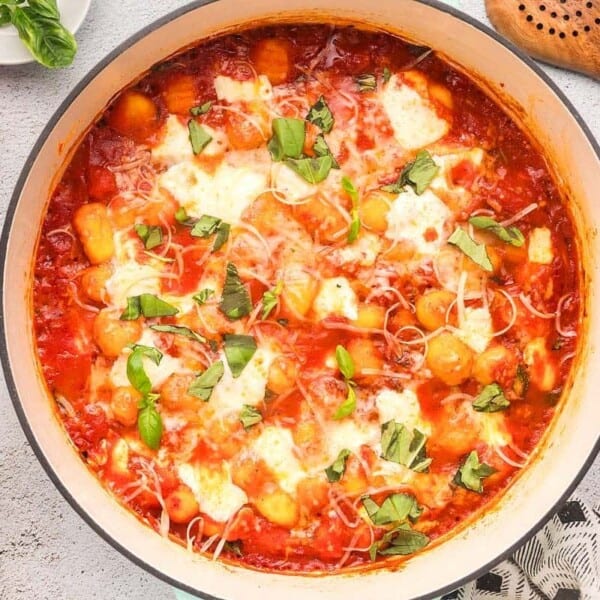 Large pot of baked gnocchi in tomato sauce with melted cheese and fresh basil, on a light surface with spoon, cheese, and oil.