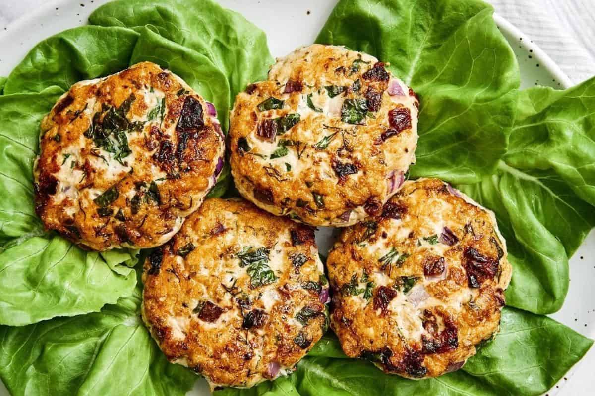 Four cooked turkey burgers, garnished with herbs and diced vegetables, are arranged on top of a bed of fresh green lettuce leaves on a white plate. The burgers appear juicy and well-seasoned, suggesting a flavorful and healthy meal.