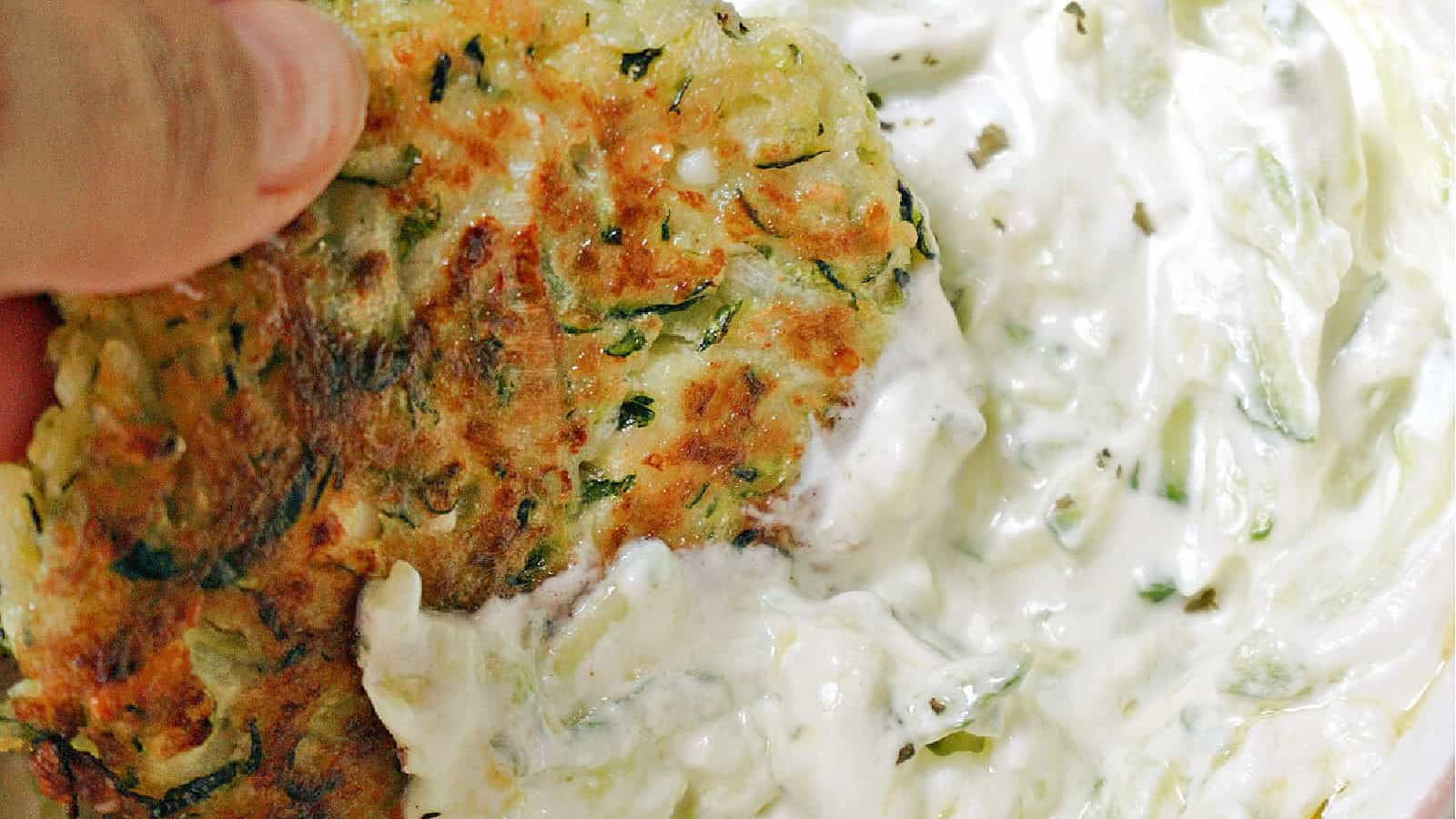 A hand-held zucchini fritter being dipped into tzatziki.