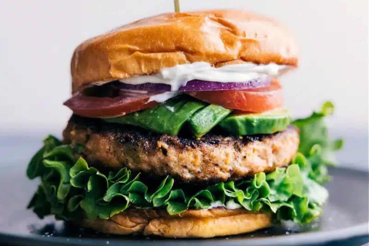 A close-up of a hamburger with a juicy grilled patty, fresh green lettuce, sliced avocado, tomato, red onion, and a layer of creamy sauce, all sandwiched between a toasted bun. A toothpick holds the burger together. The sandwich is on a dark plate.