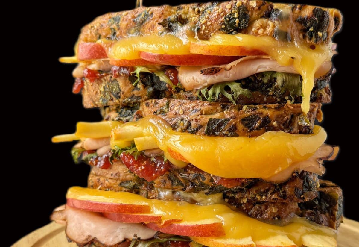 Close-up of a stacked sandwich with melted cheese, apple slices, sliced turkey, leafy greens, and multigrain bread on a wooden surface against a black background.