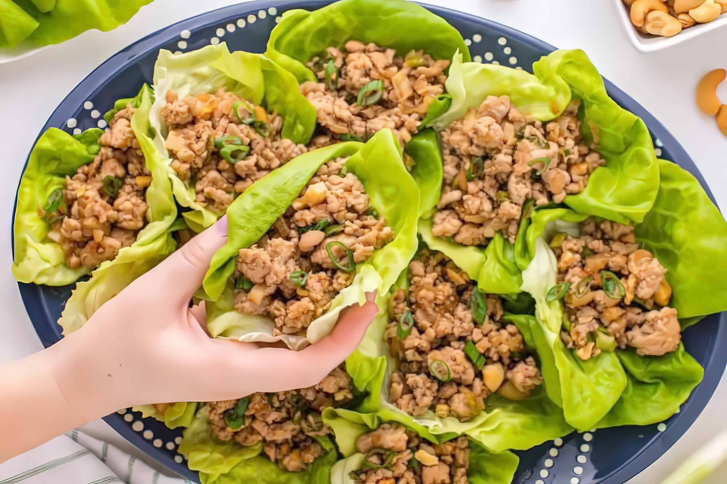 A platter is filled with lettuce wraps containing a minced chicken filling, garnished with chopped green onions. A hand is lifting one of the wraps from the platter. In the background, there are small bowls with nuts and other ingredients.