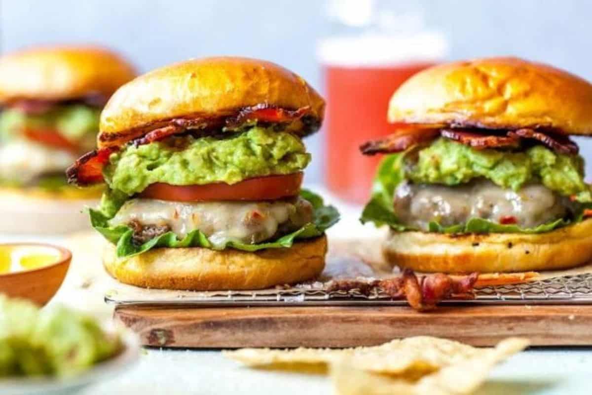 Two juicy burgers are placed on a wooden tray. Each features a toasted bun, a beef patty topped with melted cheese, fresh lettuce, a slice of tomato, a generous helping of guacamole, and crispy bacon. A drink and another burger are blurry in the background.