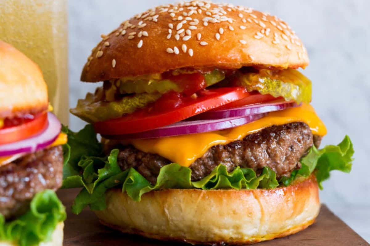 A close-up view of a cheeseburger with a sesame seed bun, featuring layers of melted cheese, a beef patty, lettuce, tomato slices, red onion rings, pickles, and ketchup. The burger is placed on a wooden surface, accompanied by a blurred background.