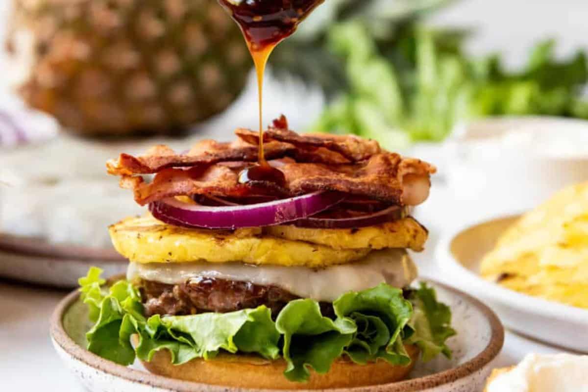 A delicious burger with a beef patty, lettuce, cheese, grilled pineapple slice, red onions, and crispy bacon is being drizzled with a thick sauce. The burger is on a ceramic plate, and fresh ingredients are visible in the background.