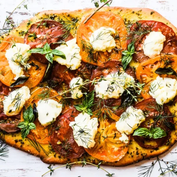 Flatbread with sliced tomatoes, cheese dollops, fresh herbs, and olive oil on a white wooden surface.