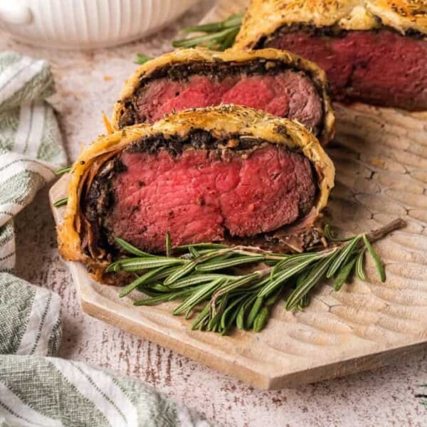 Sliced beef Wellington with a golden crust, pink center, mushroom filling on a wooden board, garnished with fresh rosemary.