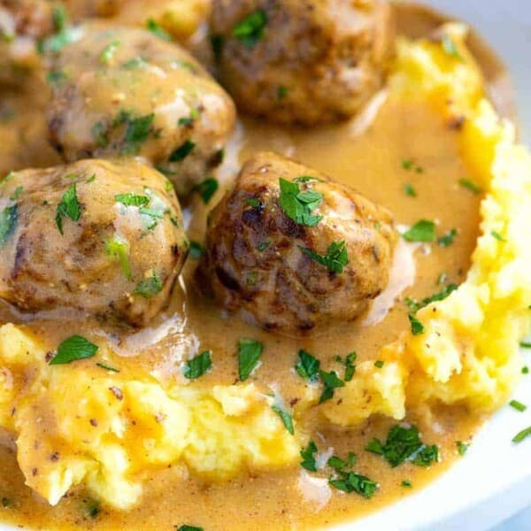 A close-up of Swedish meatballs served over creamy mashed potatoes, garnished with a generous drizzle of rich brown gravy and sprinkled with chopped parsley. The dish is presented on a white plate.