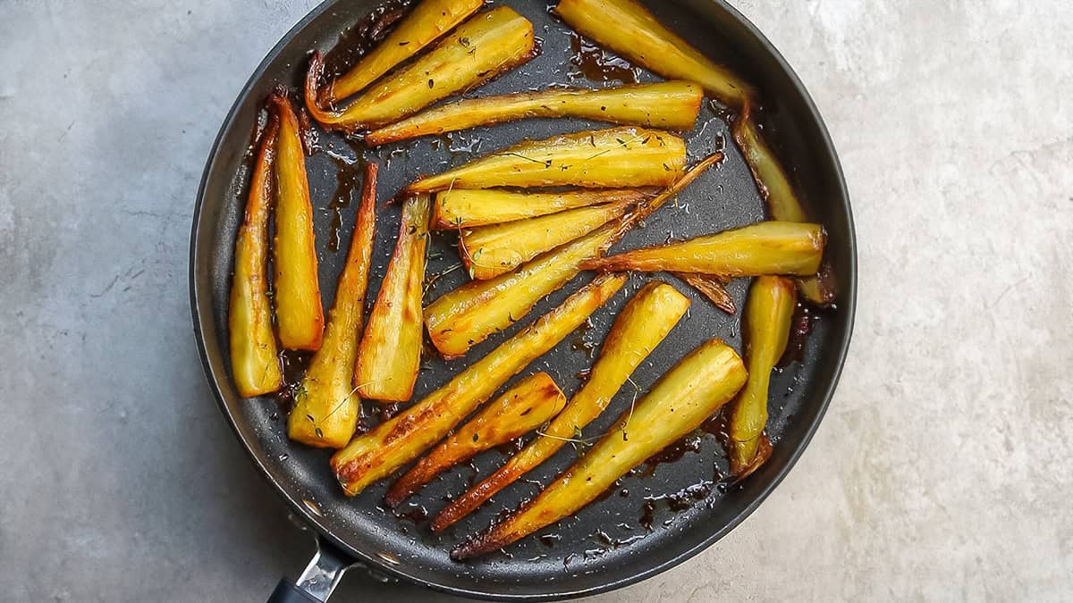 A frying pan contains several caramelized parsnip wedges, evenly spread and cooked to a golden brown with slightly charred edges, resting on a textured light gray surface.