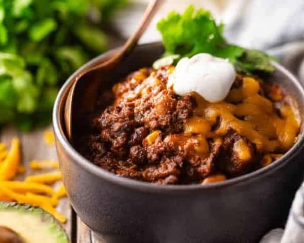 A bowl filled with savory chili topped with melted cheese, a dollop of sour cream, and a sprig of cilantro. A wooden spoon rests on the edge of the bowl. In the background, there are chopped cilantro and shredded cheese. An avocado half is visible in the foreground.