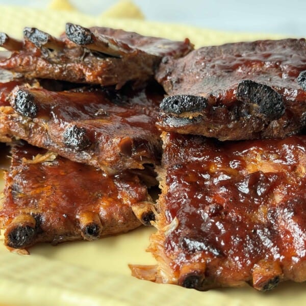 Close-up of a plate filled with barbeque ribs, covered in a rich, dark sauce with slightly charred edges. The ribs are stacked neatly on a light-colored dish, highlighting their juicy and glazed texture.