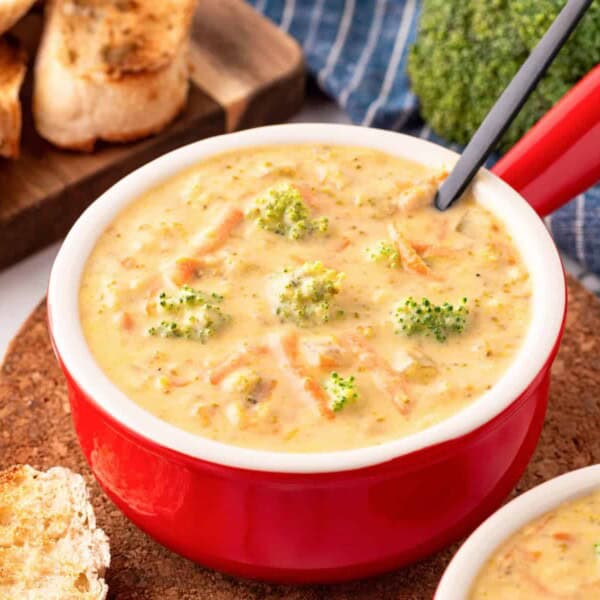 A red bowl of broccoli cheddar soup with broccoli florets sits on a cork mat next to slices of toasted bread.
