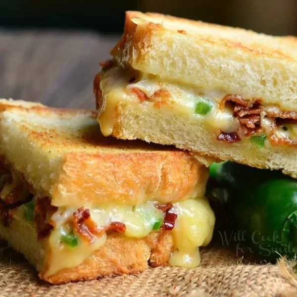 A grilled cheese sandwich with melted cheese, bacon, and jalapenos on a rustic cloth, accompanied by fresh jalapenos beside it.