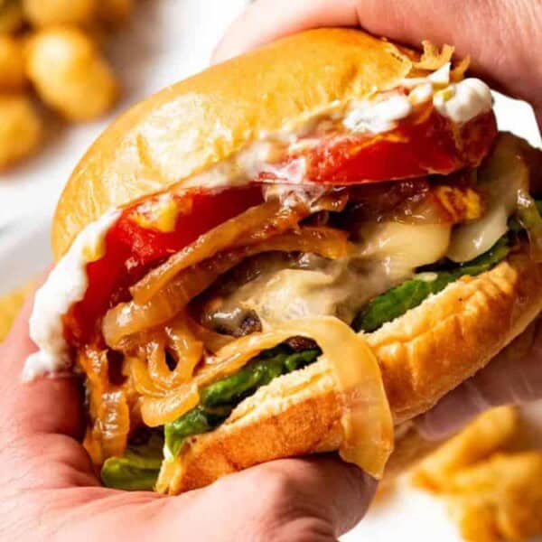 A close-up of a person holding a burger with melted cheese, caramelized onions, tomato slices, lettuce, and a generous amount of sauce. In the background, there is a plate of tater tots.