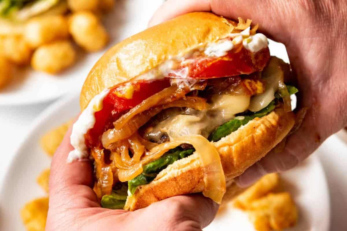 A close-up of a person holding a burger with melted cheese, caramelized onions, tomato slices, lettuce, and a generous amount of sauce. In the background, there is a plate of tater tots.