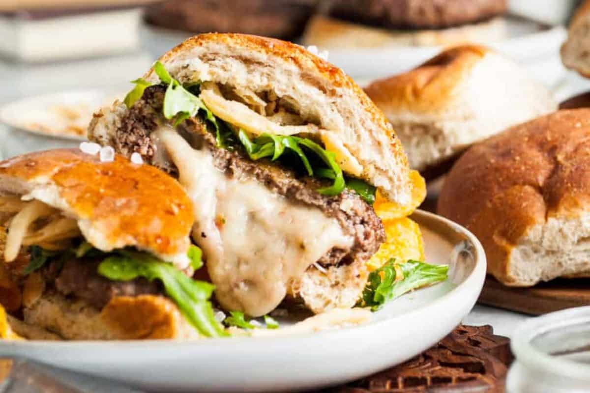 A juicy burger cut in half, placed on a white plate. The burger has a melted cheese filling, green lettuce, and fried onions inside a toasted bun. There are additional buns in the background on a wooden surface.