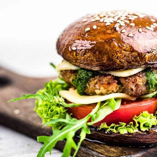A burger with a large portobello mushroom cap as the bun, filled with lettuce, tomato, cheese, patty, greens, and sauce on a wooden serving board.