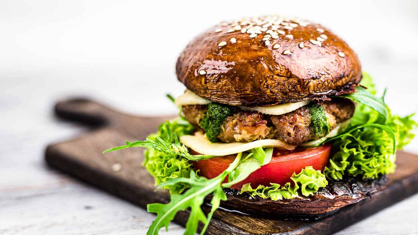 A gourmet burger with a portobello mushroom bun, grilled patty, cheese, tomato slice, fresh lettuce, arugula, and pesto sauce. The burger is placed on a wooden board with a light-colored background.
